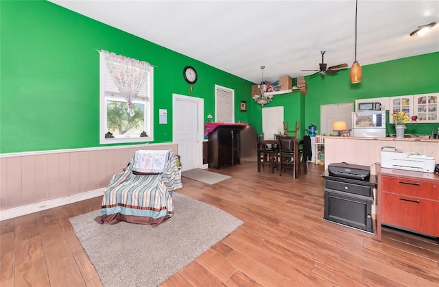 living area featuring ceiling fan, light wood-style flooring, a wainscoted wall, and a chandelier
