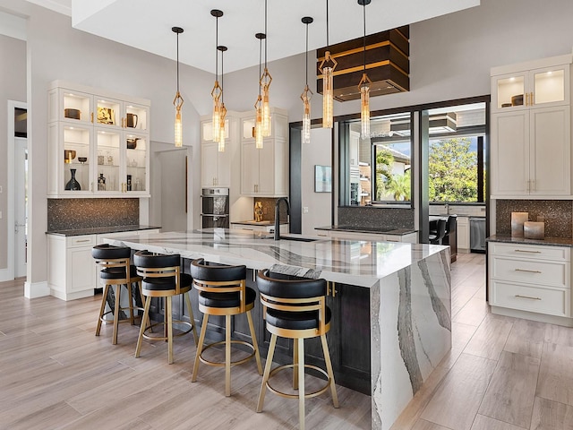 kitchen featuring double oven, tasteful backsplash, a sink, a large island with sink, and light wood finished floors
