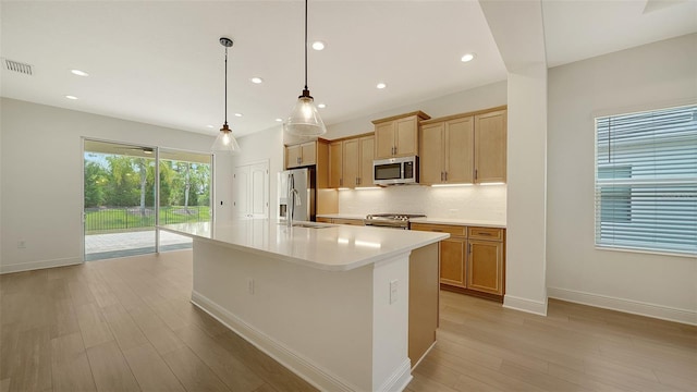 kitchen featuring appliances with stainless steel finishes, light wood-style floors, decorative backsplash, a center island with sink, and light countertops