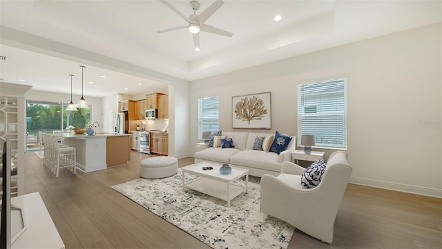 living room featuring a ceiling fan, light wood-style floors, a tray ceiling, recessed lighting, and baseboards