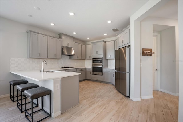 kitchen featuring under cabinet range hood, stainless steel appliances, a sink, gray cabinetry, and a peninsula
