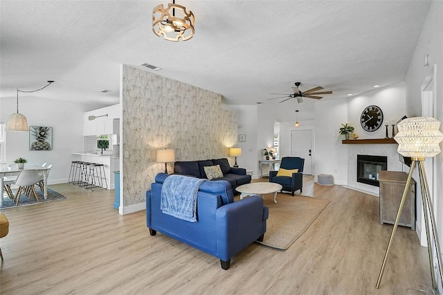 living room with light wood finished floors, a glass covered fireplace, ceiling fan, baseboards, and a textured ceiling