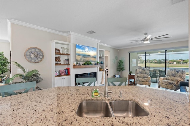 14309 Carolina Sky Pl, Lakewood Ranch, FL 34211 - kitchen with light stone countertops, a sink, ceiling fan, a glass covered fireplace, and crown molding