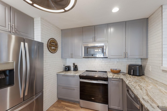 kitchen with gray cabinets, decorative backsplash, appliances with stainless steel finishes, light stone countertops, and light wood-style floors