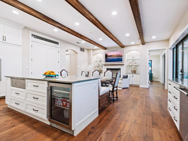 kitchen featuring beam ceiling, a center island with sink, white cabinetry, arched walkways, and beverage cooler