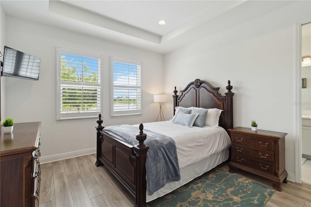 bedroom featuring light wood finished floors, a raised ceiling, baseboards, recessed lighting, and ensuite bath