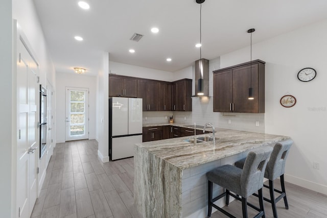 kitchen with a sink, visible vents, a peninsula, wall chimney range hood, and freestanding refrigerator