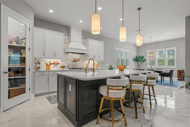 13319 Deep Blue Pl, Bradenton, FL 34211 - kitchen featuring custom range hood, light marble finish floors, light countertops, backsplash, and a kitchen island with sink