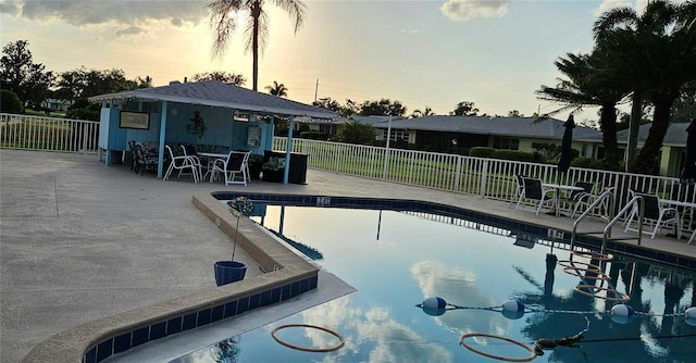 pool at dusk with an outbuilding, fence, a storage structure, a patio area, and a community pool