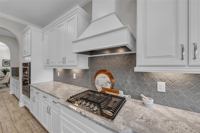 kitchen with appliances with stainless steel finishes, arched walkways, white cabinetry, custom range hood, and backsplash