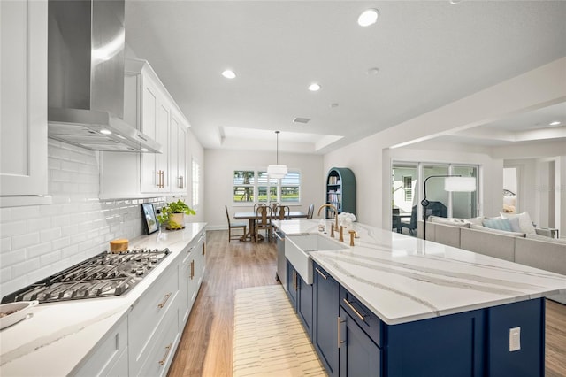 kitchen featuring wall chimney range hood, a tray ceiling, a sink, and blue cabinetry