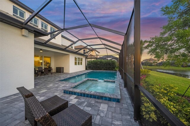 pool at dusk with a lanai, a water view, a pool with connected hot tub, and a patio area