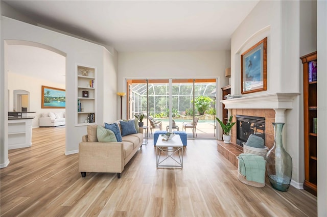living area featuring arched walkways, light wood-style flooring, built in features, a tile fireplace, and baseboards