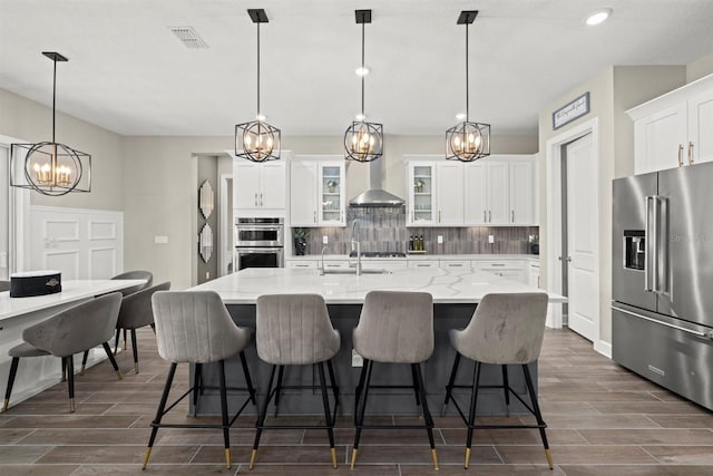 kitchen featuring decorative backsplash, wood finish floors, wall chimney range hood, a chandelier, and appliances with stainless steel finishes