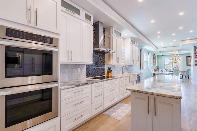 kitchen with wall chimney range hood, a center island, stainless steel double oven, white cabinetry, and a sink