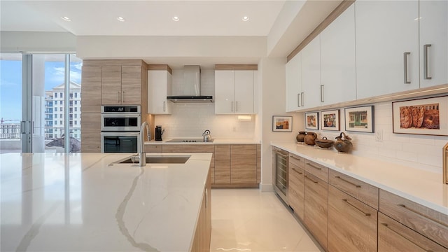 kitchen with wall chimney range hood, modern cabinets, double oven, a sink, and backsplash
