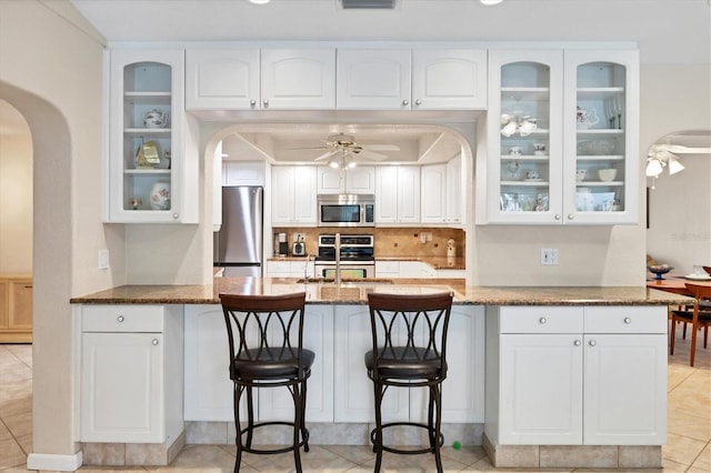 kitchen with stainless steel appliances, arched walkways, a ceiling fan, dark stone countertops, and a peninsula