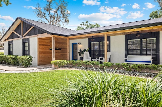 view of front of home with a front yard, stucco siding, a ceiling fan, a shingled roof, and a porch