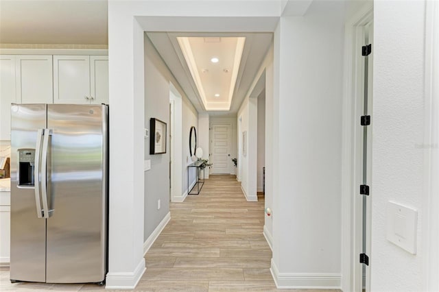 corridor featuring baseboards, a tray ceiling, and light wood-style flooring
