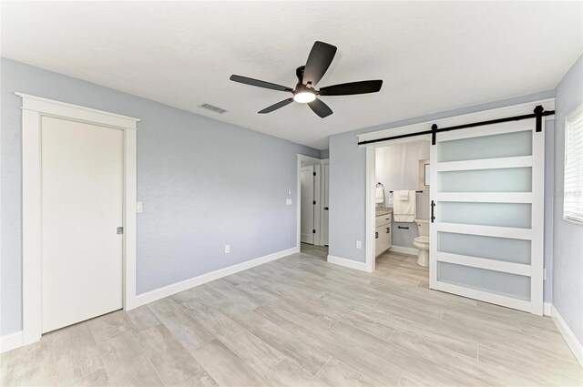 unfurnished bedroom featuring visible vents, light wood-style floors, ensuite bathroom, baseboards, and a barn door