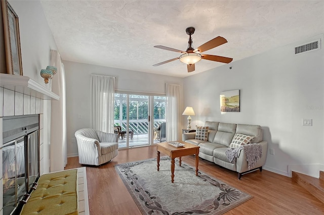living area featuring a tiled fireplace, a ceiling fan, wood finished floors, a textured ceiling, and baseboards
