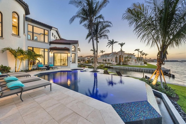 pool at dusk featuring an infinity pool, a patio area, and a water view