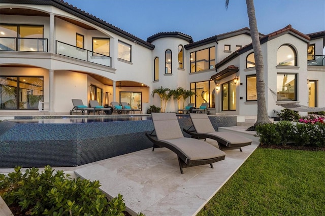 rear view of property featuring a balcony, a patio, an infinity pool, stucco siding, and a tile roof