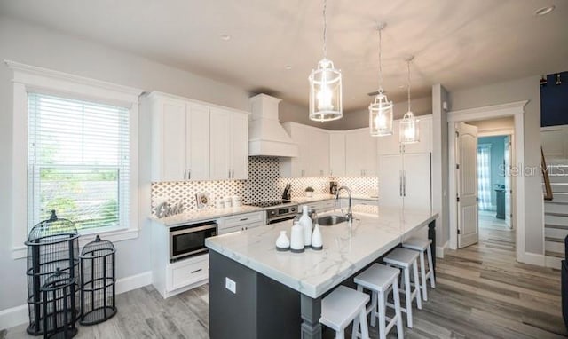kitchen with a sink, stainless steel microwave, custom exhaust hood, light wood finished floors, and tasteful backsplash