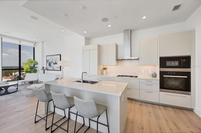 kitchen with black appliances, wall chimney exhaust hood, a sink, light wood-type flooring, and backsplash