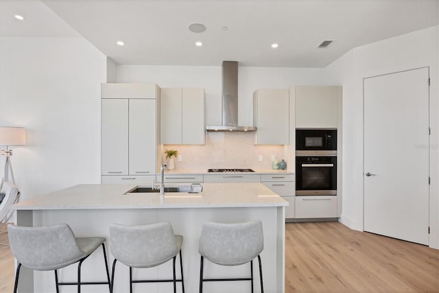 kitchen with black appliances, wall chimney exhaust hood, a sink, a breakfast bar area, and light wood-type flooring