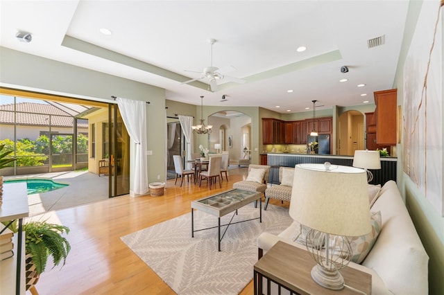 living room featuring visible vents, light wood-type flooring, a raised ceiling, arched walkways, and ceiling fan with notable chandelier