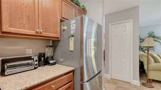 kitchen featuring freestanding refrigerator, a toaster, light stone counters, light tile patterned flooring, and brown cabinets