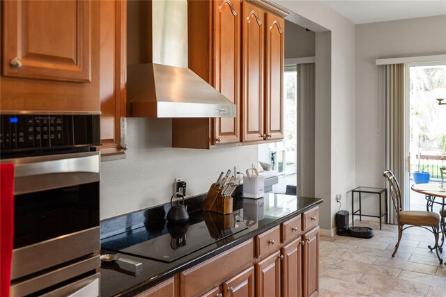 kitchen featuring wall chimney exhaust hood, stainless steel oven, black electric stovetop, brown cabinetry, and dark stone countertops