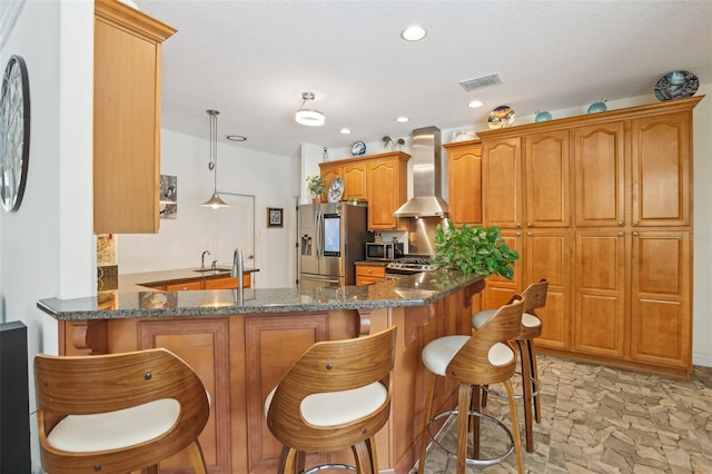 kitchen featuring brown cabinets, wall chimney exhaust hood, a kitchen breakfast bar, visible vents, and appliances with stainless steel finishes