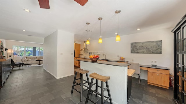 kitchen featuring wall chimney exhaust hood, dark countertops, a kitchen island, ceiling fan, and a sink