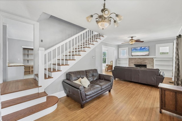 living area featuring stairs, light wood finished floors, a chandelier, a brick fireplace, and ceiling fan