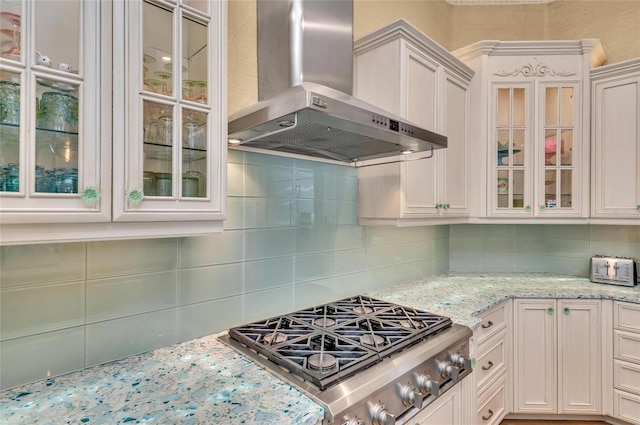 kitchen featuring wall chimney exhaust hood, stainless steel gas cooktop, decorative backsplash, light stone counters, and glass insert cabinets