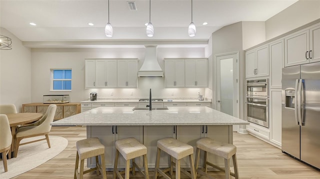 kitchen featuring stainless steel appliances, custom range hood, light wood-style flooring, a kitchen breakfast bar, and a center island with sink