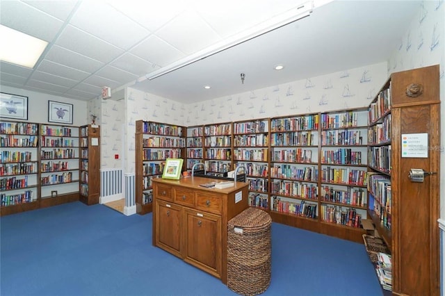 home office featuring bookshelves, wallpapered walls, carpet flooring, and a drop ceiling
