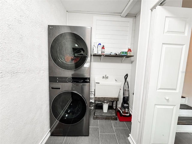 clothes washing area featuring stacked washer / drying machine, a textured wall, baseboards, a sink, and dark tile patterned floors
