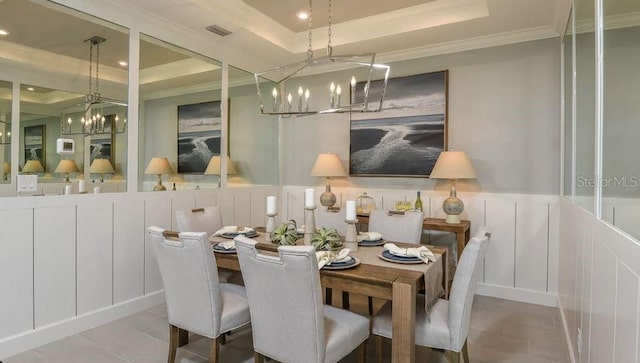 dining space featuring a raised ceiling, a chandelier, a decorative wall, crown molding, and tile patterned floors
