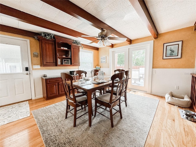 dining area featuring beam ceiling, a wainscoted wall, light wood finished floors, ceiling fan, and french doors