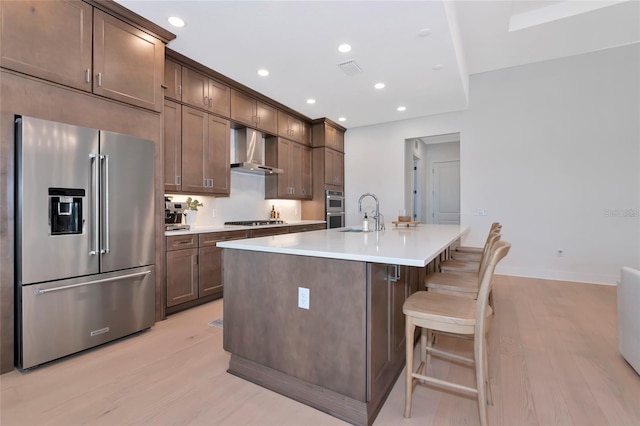 kitchen featuring appliances with stainless steel finishes, wall chimney range hood, a sink, light wood-style floors, and an island with sink