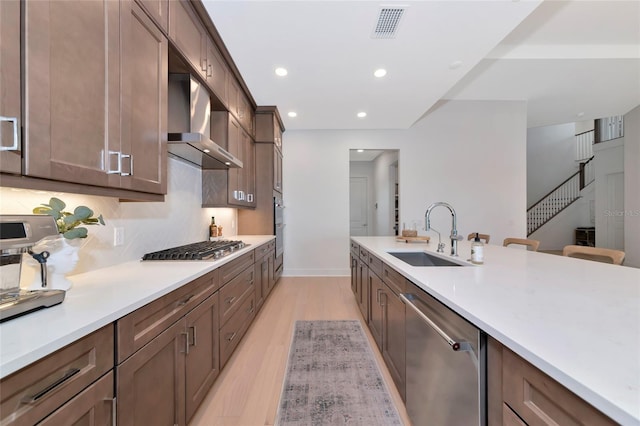 kitchen featuring appliances with stainless steel finishes, a sink, light wood-style floors, wall chimney range hood, and light countertops