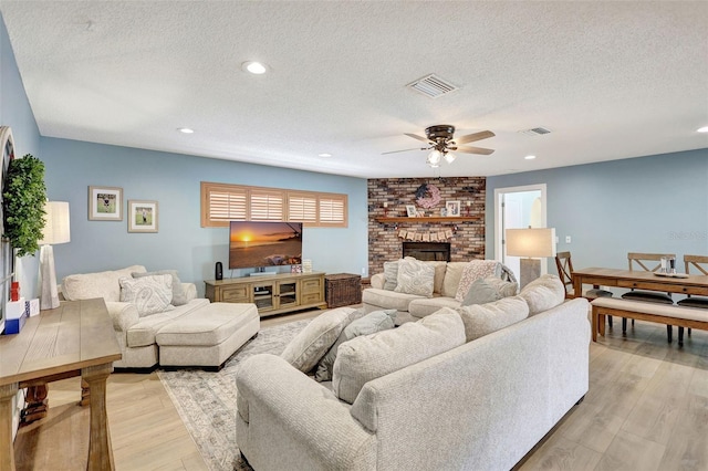 living area with ceiling fan, a textured ceiling, light wood-type flooring, a brick fireplace, and recessed lighting