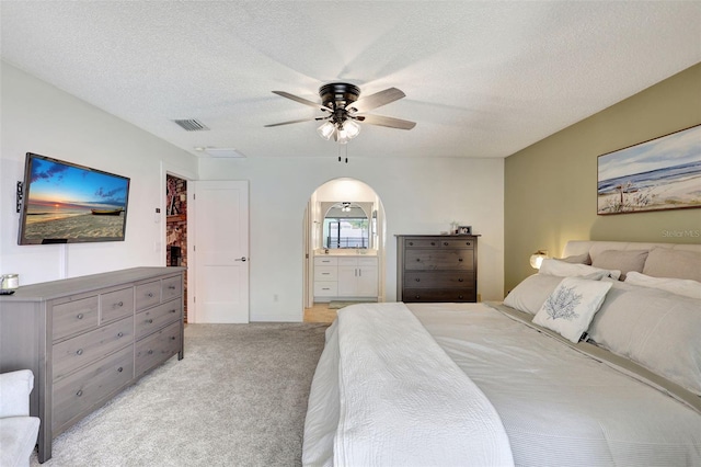 bedroom featuring light carpet, arched walkways, ensuite bath, a ceiling fan, and a textured ceiling