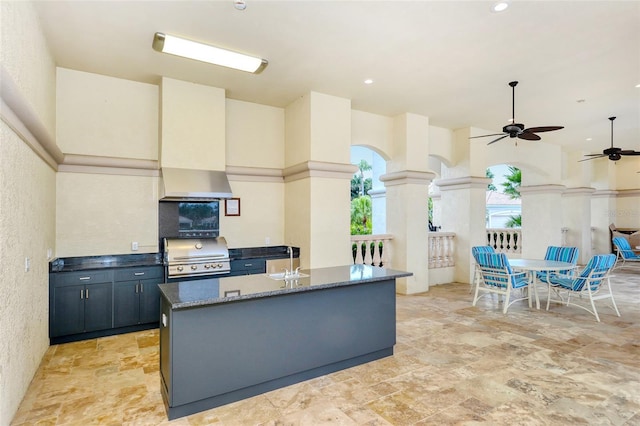 kitchen with dark stone countertops, wall chimney range hood, a ceiling fan, a textured wall, and light stone finish floors