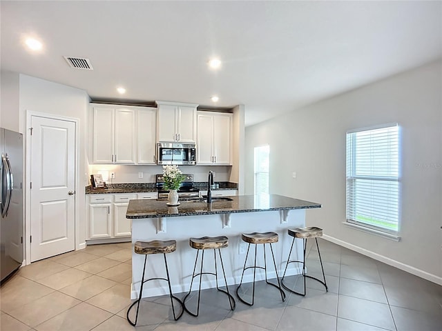 kitchen with stainless steel appliances, dark stone counters, light tile patterned floors, a kitchen island with sink, and recessed lighting
