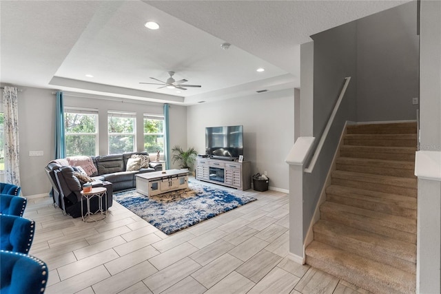 living area with a raised ceiling, ceiling fan, stairway, baseboards, and recessed lighting
