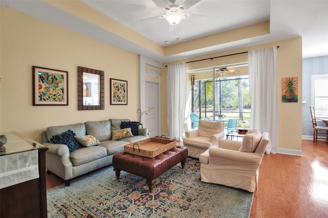 living area featuring ceiling fan, wood finished floors, baseboards, and a tray ceiling